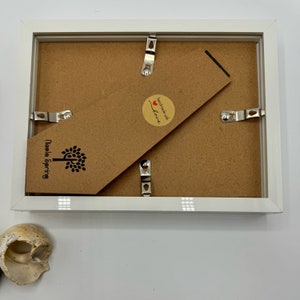 K&ouml;nnte beinhalten: Wei&szlig;er Bilderrahmen mit braunem Kartonr&uuml;cken. Der R&uuml;cken hat einen Aufkleber mit der Aufschrift "Handgefertigt mit Liebe" und ein Logo mit der Aufschrift "Needle Spring".