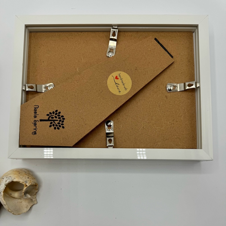 K&ouml;nnte beinhalten: Wei&szlig;er Bilderrahmen mit braunem Kartonr&uuml;cken. Der R&uuml;cken hat einen Aufkleber mit der Aufschrift "Handgefertigt mit Liebe" und ein Logo mit der Aufschrift "Needle Spring".