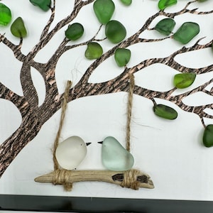K&ouml;nnte beinhalten: Ein wei&szlig;er und ein hellblauer Glasvogel auf einer Zweig-Schaukel, die an einem Ast aus bemaltem Holz mit gr&uuml;nen Glasbl&auml;ttern h&auml;ngt.