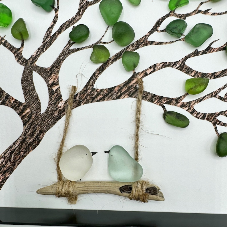 K&ouml;nnte beinhalten: Ein wei&szlig;er und ein hellblauer Glasvogel auf einer Zweig-Schaukel, die an einem Ast aus bemaltem Holz mit gr&uuml;nen Glasbl&auml;ttern h&auml;ngt.