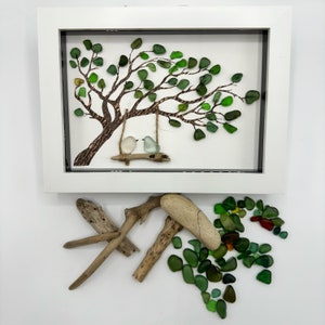K&ouml;nnte beinhalten: Ein gerahmtes Kunstwerk mit einem Baum aus gr&uuml;nem Meerglas, auf dem zwei wei&szlig;e V&ouml;gel auf einer Schaukel sitzen. Der Rahmen ist wei&szlig; und der Hintergrund ist wei&szlig;. Um den Rahmen herum liegen St&uuml;cke Treibholz und Meerglas.