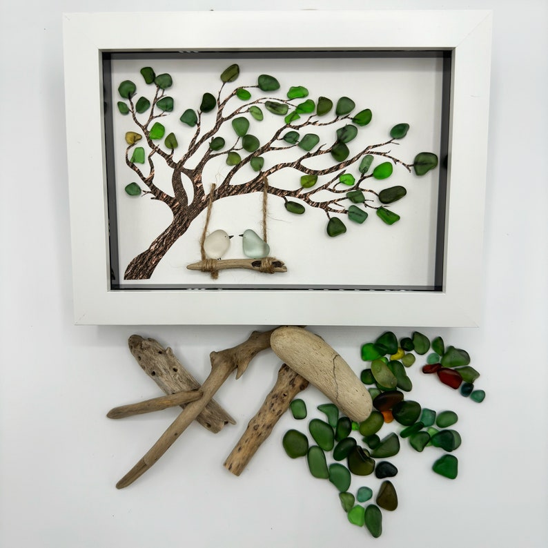 K&ouml;nnte beinhalten: Ein gerahmtes Kunstwerk mit einem Baum aus gr&uuml;nem Meerglas, auf dem zwei wei&szlig;e V&ouml;gel auf einer Schaukel sitzen. Der Rahmen ist wei&szlig; und der Hintergrund ist wei&szlig;. Um den Rahmen herum liegen St&uuml;cke Treibholz und Meerglas.
