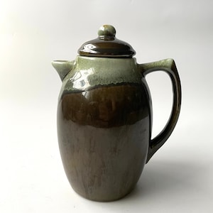 May include: A ceramic teapot with a lid, featuring a dark brown base that fades into a lighter green at the top. The teapot has a handle and a spout, and is set against a white background.