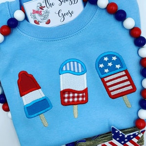 May include: A light blue sweatshirt with three embroidered popsicles. The popsicles are red, white, and blue and have a gingham pattern, stripes, and stars. The text "The Snazzy Goose" is on a white tag.