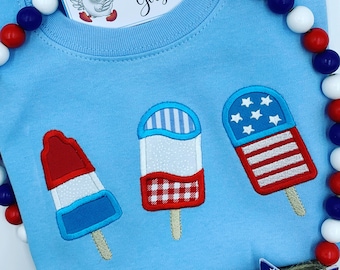 May include: A light blue sweatshirt with three embroidered popsicles. The popsicles are red, white, and blue and have a gingham pattern, stripes, and stars. The text "The Snazzy Goose" is on a white tag.