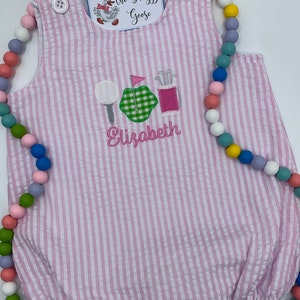 May include: Pink and white striped baby onesie with a golf-themed embroidered design. The design includes a golf ball, a golf club, a green flag, and a golf bag. The onesie is personalized with the name "Elizabeth".