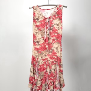 May include: A sleeveless dress with a floral pattern of red roses and brown accents on a beige background. The dress has a tie at the neckline and a ruffled hem. It hangs on a silver hanger.