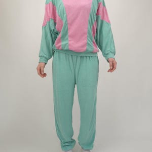 Może przedstawiać: Dwuczęściowy dres w stylu retro. Góra jest różowa i miętowa, z blokowym wzorem kolorystycznym. Pasujące spodnie są miętowe. Strój jest wykonany z miękkiej, teksturowanej tkaniny. Osoba ma na sobie białe kapcie.