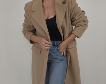 Könnte beinhalten: Ein beigefarbener Blazer mit klassischem Revers und zwei Vordertaschen. Der Blazer wird über einem schwarzen Oberteil und einer blauen Jeans getragen. Das Outfit ist schlicht und stilvoll, passend für verschiedene Anlässe.