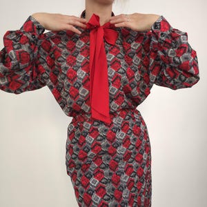 May include: A vintage dress featuring a red bow and a geometric pattern in red, gray, and black. The long-sleeved dress has a fitted waist and a knee-length skirt, perfect for a retro look.