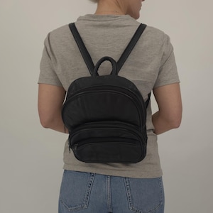 Puede incluir: Mochila de cuero negro con dos compartimentos con cremallera y correas ajustables. La mochila pequeña tiene un asa superior y se lleva en la espalda. La bolsa está hecha de un material liso y duradero.