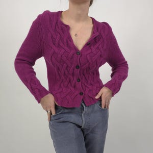 Può includere: Un cardigan magenta a trecce con scollo a V e chiusura con bottoni. Il maglione a maniche lunghe presenta un motivo strutturato ed è abbinato a jeans blu. La modella indossa una collana e anelli d'argento.