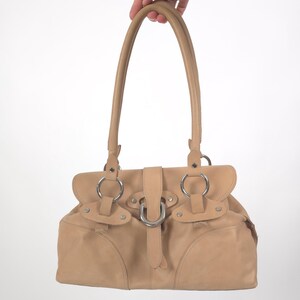 May include: A tan leather handbag with a silver buckle and two silver rings on each side. The bag has a top handle and a flap closure.