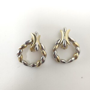 Pode incluir: Um par de brincos de pressão bicolores. Os brincos apresentam um design floral dourado na parte superior e um aro torcido prateado e dourado. Os brincos estão sobre um fundo branco.