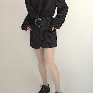 May include: Black pinstripe blazer with a wide black belt featuring a large silver buckle. The blazer is paired with white tights and black lace-up shoes. The outfit is a mix of classic and edgy styles.