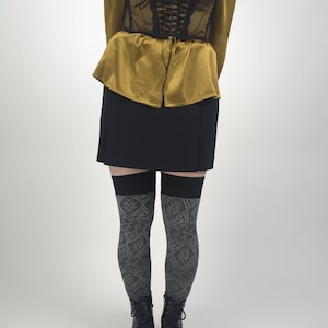 May include: A black lace corset with a lace-up front is worn over a mustard-colored blouse. A black mini-skirt and patterned thigh-high stockings are also visible. The outfit is completed with black platform boots.