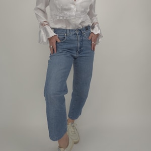 Könnte beinhalten: Eine weiße Bluse mit V-Ausschnitt und langen Ärmeln, verziert mit floraler Stickerei und Spitze. Dazu werden hellblaue Jeans und weiße Turnschuhe getragen. Das Outfit wirkt lässig und stilvoll.