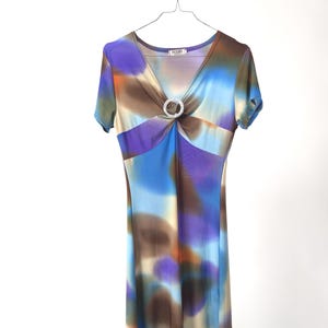 Könnte beinhalten: Kurzärmeliges Kleid mit V-Ausschnitt und dekorativem Ringdetail. Das Kleid hat ein farbenfrohes abstraktes Muster in Blau-, Lila-, Braun- und Orangetönen. Es hängt an einem Metallbügel.