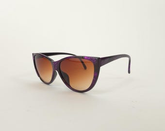 May include: Cat-eye sunglasses with a purple speckled frame and gradient brown lenses. The arms are black. These sunglasses are a stylish accessory for eye protection.
