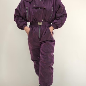 Puede incluir: Traje de esquí morado vintage con chaqueta con capucha y pantalón a juego. La chaqueta tiene múltiples bolsillos con cremallera y cinturón en la cintura. El traje es de material brillante y acolchado, y los pantalones tienen puños elásticos.