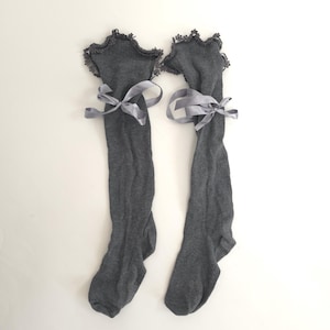 May include: A pair of gray knee-high socks with a scalloped lace trim at the top. Each sock is tied with a gray satin ribbon. The socks are displayed on a white background.