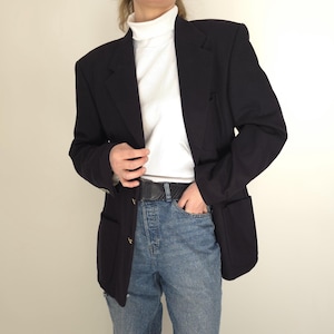 May include: A black blazer with gold buttons, paired with a white turtleneck and blue jeans. The blazer has a classic design with a tailored fit and pockets. The outfit is completed with a black belt.