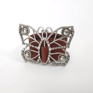 May include: A brown suede belt featuring a large, silver-toned metal buckle shaped like a butterfly. The buckle has an open-work wing design with four circular, polished silver accents positioned on the upper and lower sections of the wings.