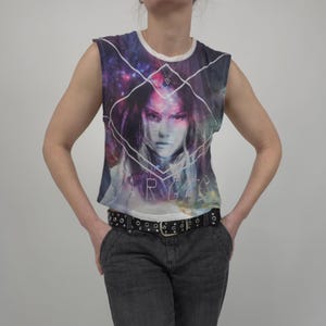May include: Sleeveless t-shirt with a galaxy-themed graphic featuring a woman's face and the word "ARGIA" in white. The shirt has a white neckline and armholes. The person is wearing a black studded belt and dark gray pants.