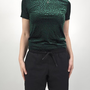 May include: A dark green velvet top with a black leopard print pattern. The top has short sleeves and a gathered waist. Paired with black drawstring pants. A silver necklace is visible.