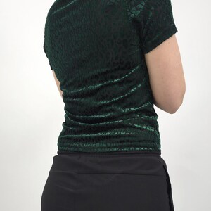 May include: A dark green, short-sleeved top with a velvet texture and a subtle animal print pattern. The top is paired with black pants. The model is facing away from the camera.