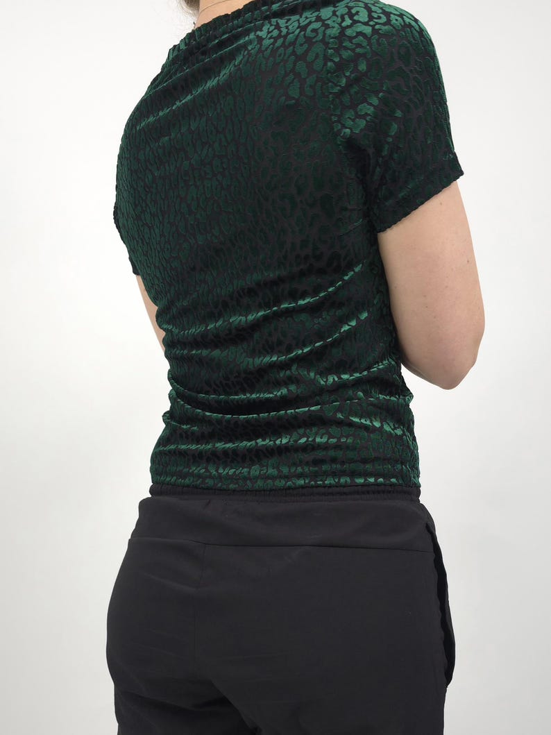 May include: A dark green, short-sleeved top with a velvet texture and a subtle animal print pattern. The top is paired with black pants. The model is facing away from the camera.