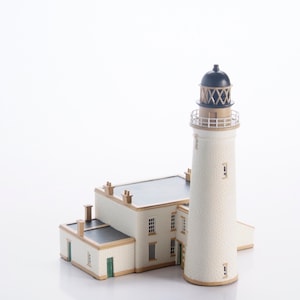 Puede incluir: Maqueta detallada de un faro. La estructura presenta un exterior blanco con un tejado negro y una alta torre cilíndrica. La torre tiene una linterna negra y dorada. El modelo incluye pequeñas ventanas y puertas.