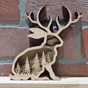 May include: A wooden cutout of a rabbit with antlers, featuring a layered design that reveals a forest scene inside. The rabbit is standing on a wooden base.