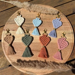 Peut inclure: Plusieurs porte-clés en macramé en forme de cœur, dans des tons crème, bleu, lavande, marron, vert, orange et rose. Chaque porte-clés est doté d'un fermoir doré et d'un pompon assorti. Les porte-clés sont disposés sur un plateau en bois.