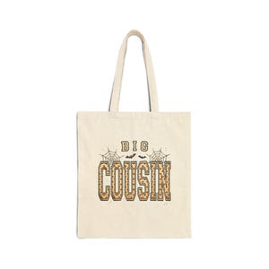 May include: A beige canvas tote bag featuring the text BIG COUSIN in a tan and orange pumpkin-patterned font. The design includes two spider webs on the sides and two small black bats centered above the word COUSIN on the front of the bag.