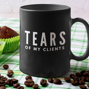 May include: A matte black ceramic mug featuring the white text "TEARS OF MY CLIENTS" printed on the front. The mug has a standard cylindrical shape with a side handle and is positioned next to three chocolate muffins and scattered coffee beans.
