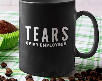 May include: A matte black ceramic mug featuring the white text "TEARS OF MY EMPLOYEES" printed on the front. The mug has a standard cylindrical shape with a curved handle on the right side, positioned next to three chocolate muffins and coffee beans.