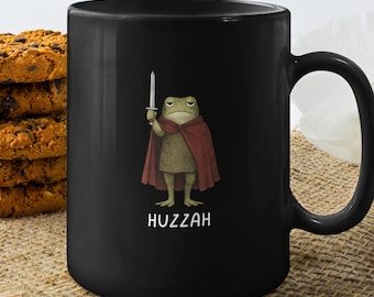 May include: Black ceramic mug featuring a cartoon frog in a red cape holding a sword. The frog has a stern expression. The word "Huzzah" is printed below the frog in white. A stack of cookies is on the left.