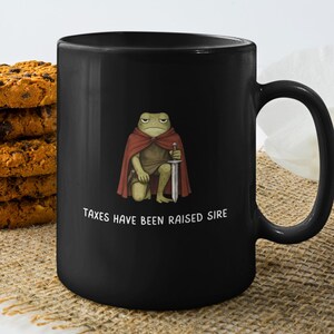 May include: Black ceramic mug featuring a cartoon frog in a red cape holding a sword. The text "TAXES HAVE BEEN RAISED SIRE" is printed below the frog. A stack of chocolate chip cookies and a white flower are in the background.