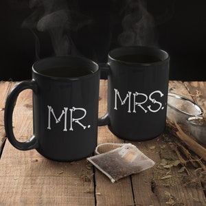 May include: Two black coffee mugs with white bone-shaped letters spelling "MR." and "MRS." on a wooden surface with a tea bag and loose tea leaves.