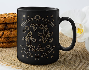 May include: Black ceramic mug featuring a gold illustration of the Pisces zodiac symbol. The design includes two fish, bubbles, and celestial elements. The mug is paired with a stack of chocolate chip cookies.