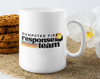 May include: White ceramic mug with the text "DUMPSTER FIRE response team" in black, with a graphic of a burning dumpster. A stack of chocolate chip cookies and a white flower are in the background.