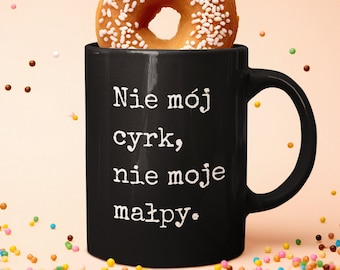 May include: Black coffee mug with white text that reads "Nie mój cyrk, nie moje małpy." A donut with white sprinkles is sitting on top of the mug. Colorful sprinkles are scattered around the mug.