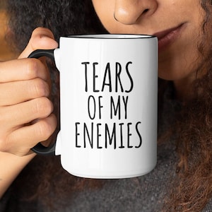 May include: A white ceramic mug with a black rim and handle. The mug has black text that reads "Tears of my Enemies".