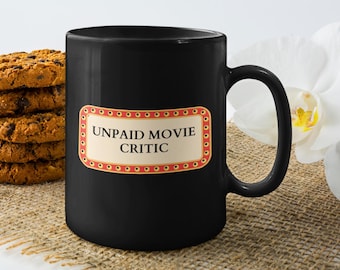 May include: Black coffee mug with the text "UNPAID MOVIE CRITIC" in a vintage-style frame. A stack of chocolate chip cookies sits to the left of the mug. A white orchid flower is in the background.