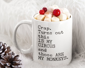 May include: White ceramic mug filled with marshmallows and topped with red berries. The mug has the text "Crap. Turns out this IS MY CIRCUS and those ARE MY MONKEYS." A pinecone is next to the mug.