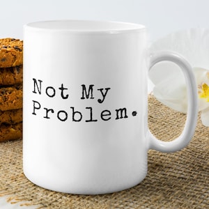 May include: White ceramic mug with the text "Not My Problem" in black. A stack of chocolate chip cookies and white orchid flowers are in the background. The mug has a curved handle.