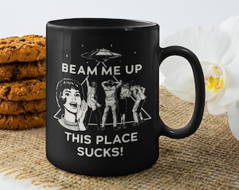 May include: A black ceramic mug featuring a white graphic print of a flying saucer above a group of people and the text "BEAM ME UP THIS PLACE SUCKS!" The design includes a triangular outline and a person looking up with an open mouth.