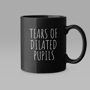 May include: A black ceramic mug featuring the white text "TEARS OF DILATED PUPILS" printed in a clean, sans-serif font. The mug has a smooth, glossy finish, a cylindrical body, and a curved handle on the right side for a comfortable grip.