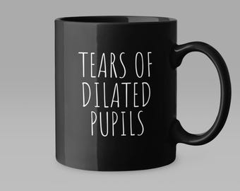 May include: Black ceramic mug with a curved handle. The mug features the white text "TEARS OF DILATED PUPILS" in a sans-serif font. The mug is set against a neutral gray background.